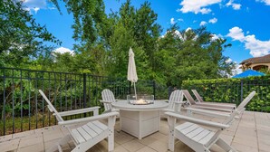 A patio with a round table and four Adirondack chairs, surrounded by a black metal fence, with trees and a building in the background. - Desert Waves at Reunion-TT489 (Kissimmee)