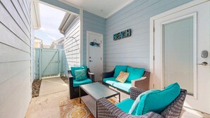 Un patio exterior cubierto con muebles de mimbre y cojines color turquesa, una puerta decorada con motivos de peces y un cartel que dice 'Beach'.
