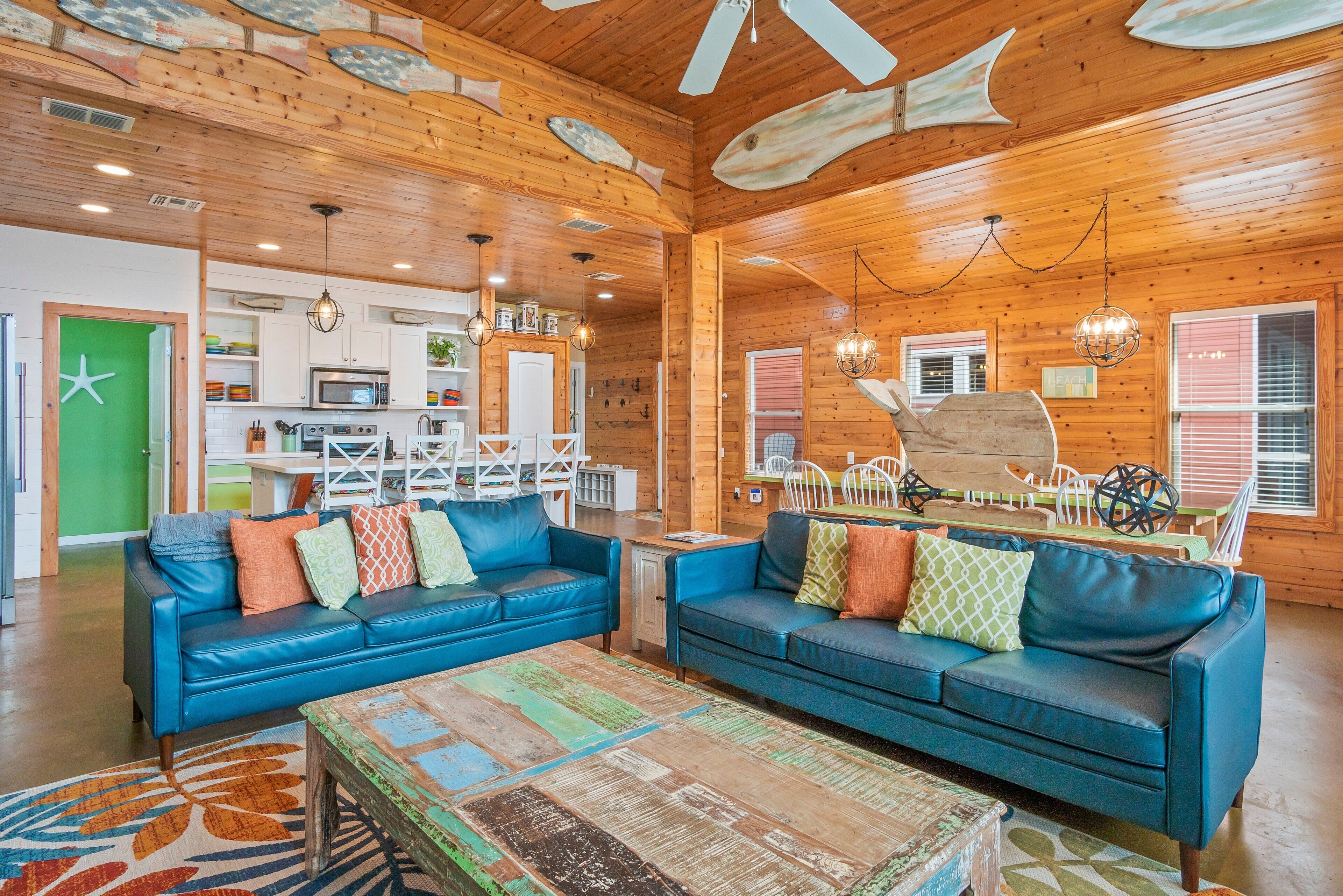 A spacious living area with two blue leather sofas, a wooden ceiling with fish decorations, and a kitchen in the background.