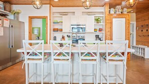 Une cuisine moderne avec des meubles blancs, un réfrigérateur en acier inoxydable, un four à micro-ondes et un évier. On y trouve quatre chaises blanches avec des coussins colorés, disposées autour d’une table en bois.
