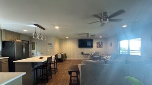 Een moderne keuken met een centraal eiland, barstoelen en een eethoek. Er is een woonkamer met een bank en een televisie aan de muur.