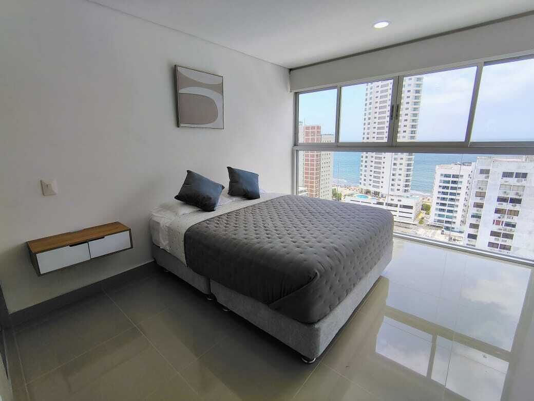 A modern bedroom with a large bed, a nightstand, a small wooden drawer unit, and a large window offering a city view.
