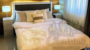 A hotel room with a large bed, two bedside lamps, a headboard with vertical slats, and a view of a cityscape through sheer curtains. - Brenthill Baguio (Baguio)