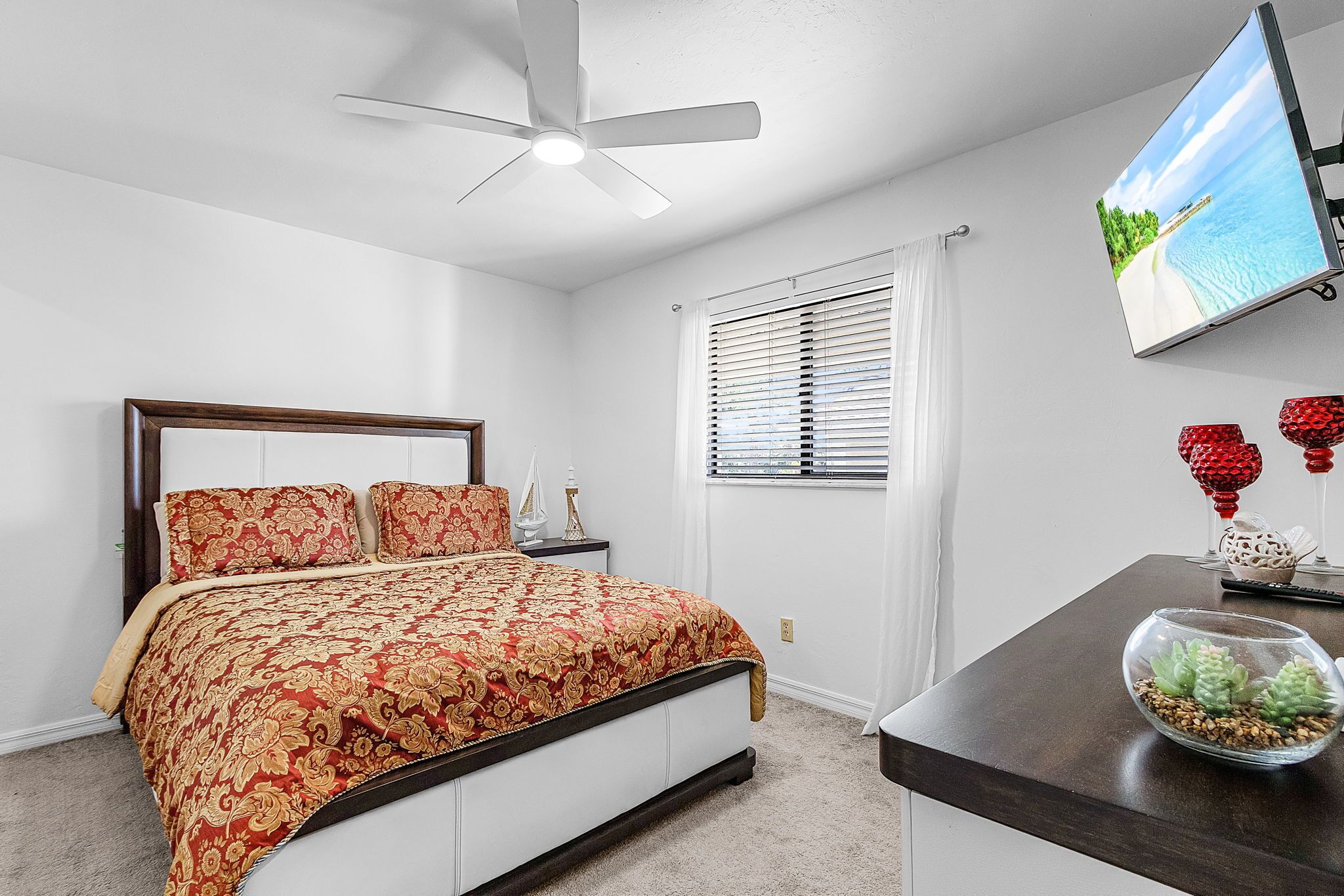 A bedroom with a bed, a ceiling fan, a TV, and a window with blinds.
