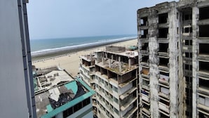 A high-rise building with multiple floors, some of which are under construction. A beach is visible in the background.