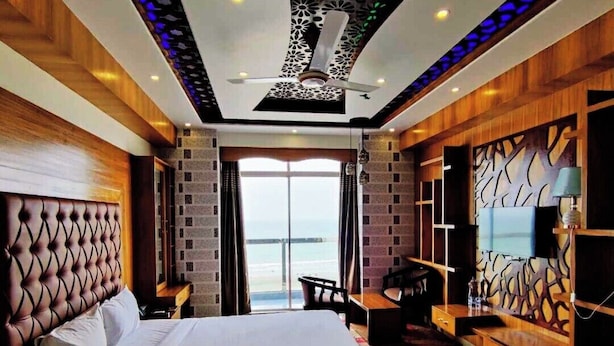 A modern hotel room with a large bed, a desk, and a TV. The room has a balcony with a view of the sea.