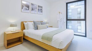 A bedroom with a wooden bed, white bedding, a nightstand with a lamp, a large window, and three framed pictures on the wall. - Cedar Vista - condo with stunning mountain views (Revelstoke)