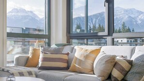 A modern living room with a grey sofa, multiple cushions, and a round wooden table. Large windows offer a view of snow-capped mountains. - Cedar Vista - condo with stunning mountain views (Revelstoke)