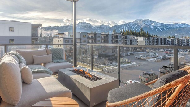 A balcony with a fire pit, outdoor seating, and a view of snow-capped mountains and modern buildings. - Cedar Vista - condo with stunning mountain views (Revelstoke)