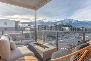 A balcony with a fire pit, outdoor seating, and a view of snow-capped mountains and modern buildings.
