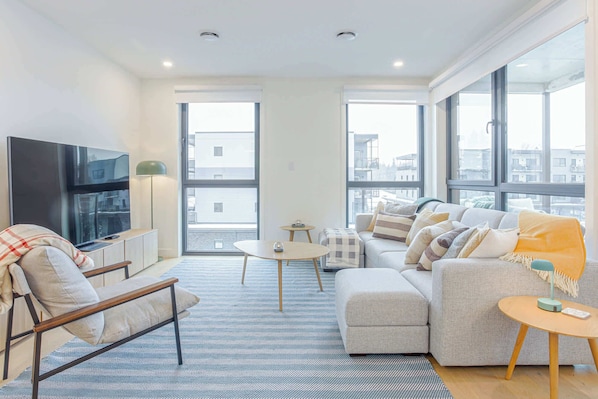 A modern living room with a sofa, armchair, TV, and a view of the city through large windows.