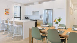 A modern kitchen with white cabinets, a stainless steel refrigerator, and a dining area with a wooden table and green chairs. - Cedar Vista - condo with stunning mountain views (Revelstoke)