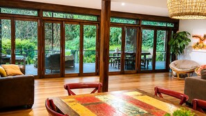 Une pièce spacieuse avec une grande table à manger en bois, des chaises rouges et une vue sur une végétation luxuriante, visible à travers de grandes portes-fenêtres coulissantes.