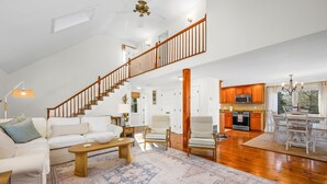 A spacious living room with a high ceiling, a wooden staircase, a kitchen with wooden cabinets, and a dining area with a chandelier. - Cozy Cape Retreat with Fenced Yard & Fire Pit (Harwich)