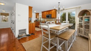 A kitchen with wooden cabinets, a central island, and a dining area with white chairs. - Cozy Cape Retreat with Fenced Yard & Fire Pit (Harwich)