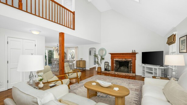 A spacious living room with a fireplace, a dining area, and a staircase leading to an upper level. - Cozy Cape Retreat with Fenced Yard & Fire Pit (Harwich)
