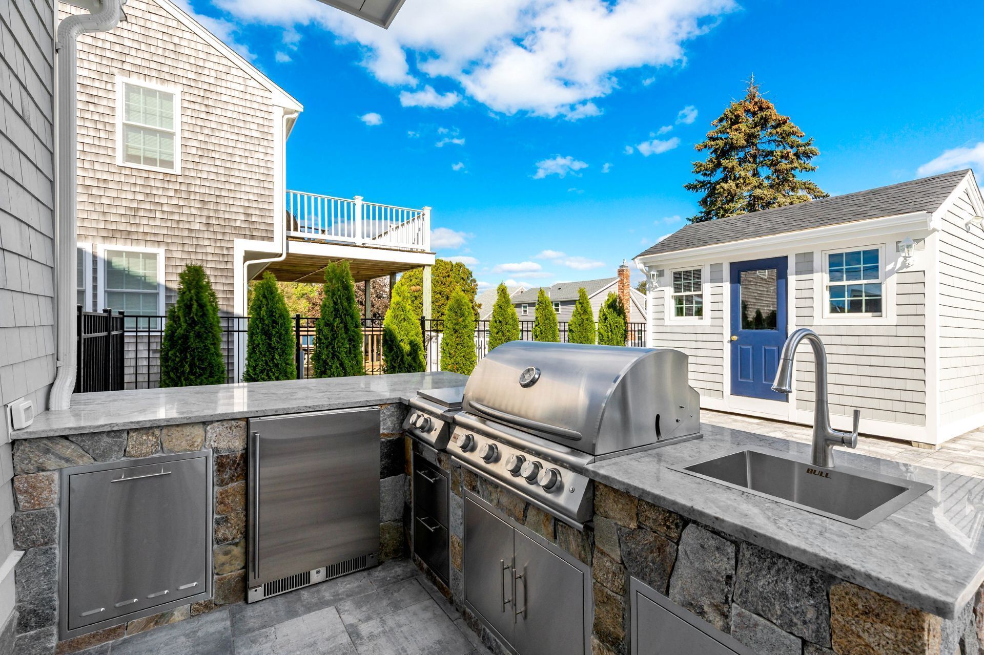 An outdoor kitchen with a grill, sink, and stone accents, set against a residential backdrop with a blue door and a tree.