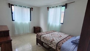 A bedroom with a bed, a wooden dresser, a small nightstand, and a window with a curtain.