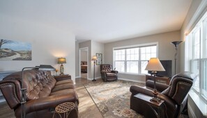 A living room with a brown leather sofa, a patterned rug, and a large window providing natural light. - Peaceful 3 Bedroom house with 157 Acres to explore. Pet Friendly. Fountain, MI. (Fountain)