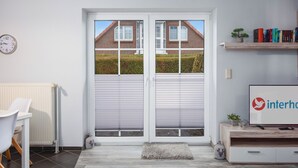 Un moderno salón con una puerta corrediza de vidrio que da a un jardín, un radiador blanco, una silla blanca, una pequeña mesa, una estantería con libros y un televisor sobre un mueble de madera.
