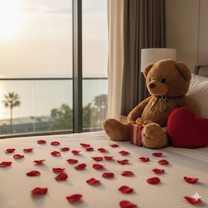 Een teddybeer op een bed met rozenblaadjes en een hart, bij een raam met uitzicht.