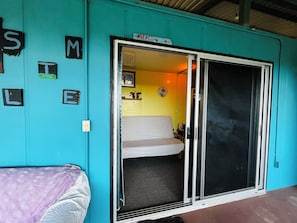 A teal-colored room with a sliding glass door leading to a yellow interior. There is a bed with a patterned cover and a chair with a purple cover. - Amazing Ocean view guest house, just next to a monkey trail (Guanacaste Province)