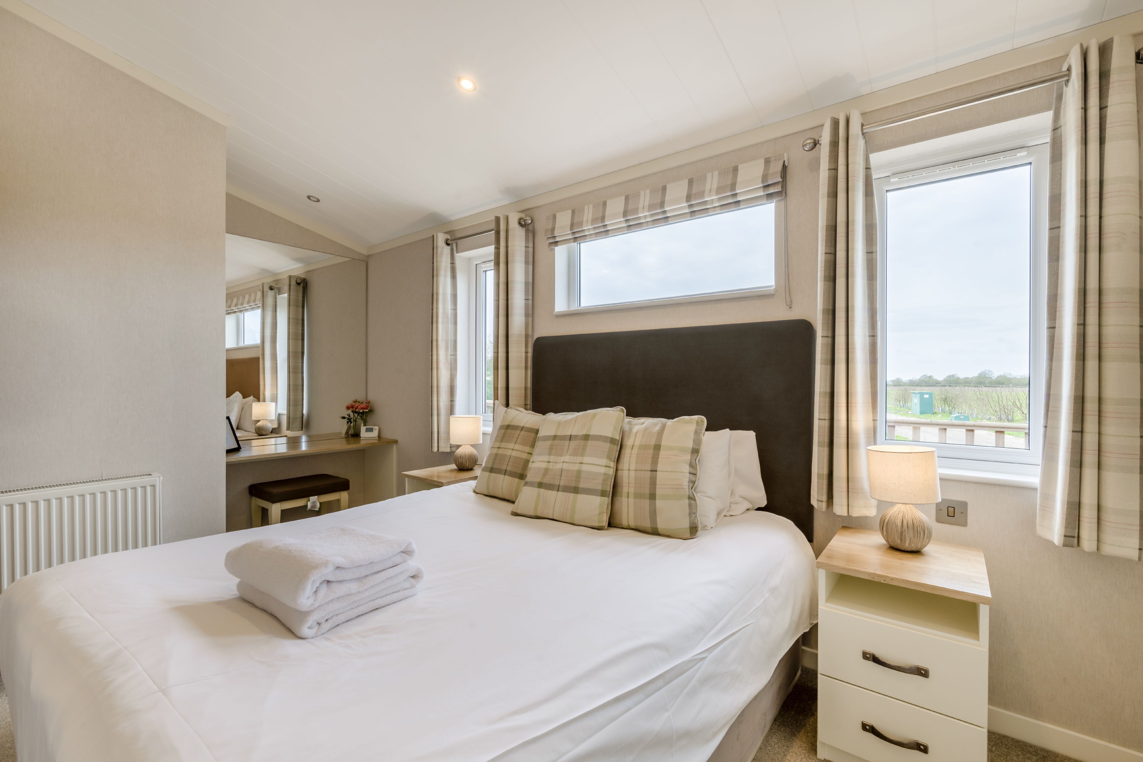 Una cama bien hecha con almohadas, una mesita de noche con una lámpara, una ventana con cortinas y un espejo en la pared.