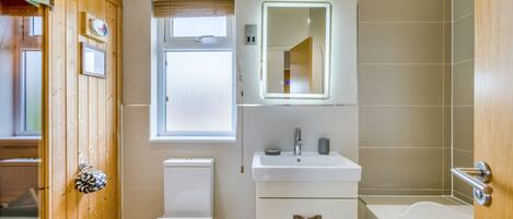 Et badeværelse med dør i træ, toilet, vask med spejl og badekar.