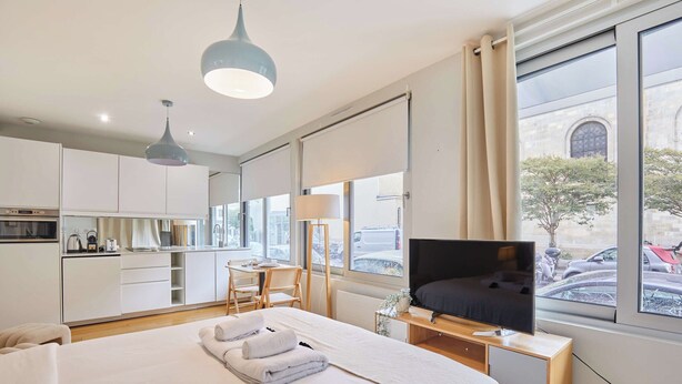 A modern hotel room with a large bed, a TV, a desk, and a view of the street through large windows. - Modern Suite Invalides Tour Eiffel (Paris)