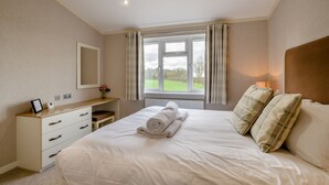 Een slaapkamer met een bed, nachtkastje, spiegel, raam met gordijnen en uitzicht op groen.
