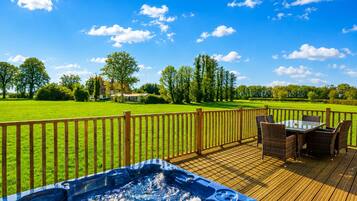 A wooden deck with a hot tub and outdoor furniture, overlooking a grassy field and trees.