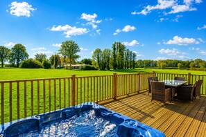 Una terraza de madera con un jacuzzi y mobiliario exterior, con vistas a un campo cubierto de césped y árboles.