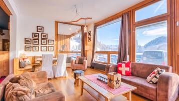 Un salon chaleureux avec un plancher en bois, un canapé, une table basse et une cheminée. Les murs sont ornés de tableaux encadrés, et une fenêtre offre une vue sur les montagnes.