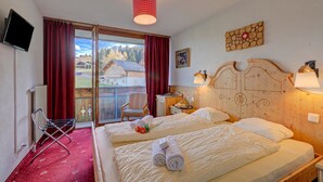 Habitació d'hotel amb dos llits, televisió, cadira, panell de capçalera de fusta i vista a una casa per una porta de vidre.
