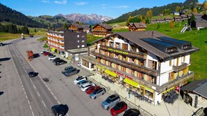 Un hotel amb un balcó destacat, un pàrquing amb diversos cotxes i una muntanya de fons.