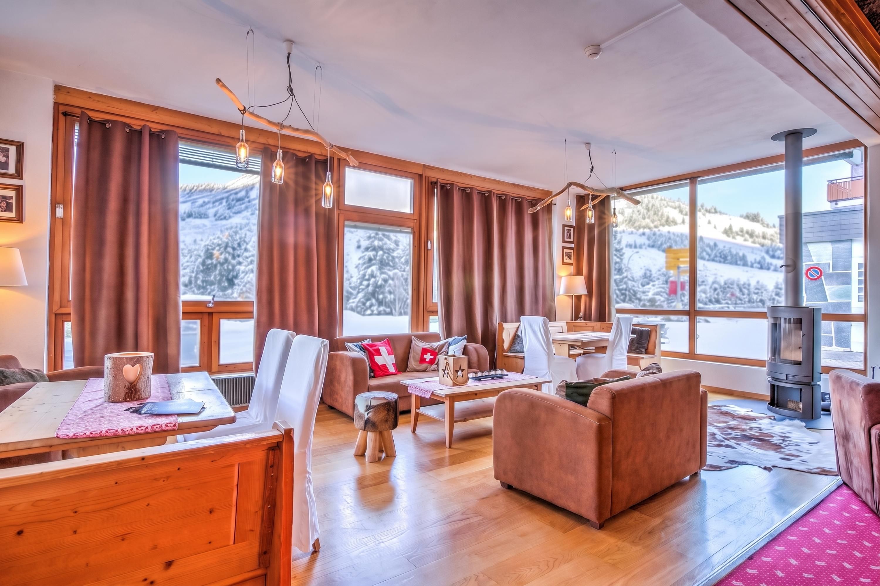 Ein gemütliches Wohnzimmer mit Kamin, Holzmöbeln und großen Fenstern mit Blick auf verschneite Berge.