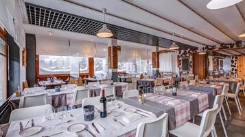 Ein Restaurant mit mehreren gedeckten Tischen, das modernes Interieur mit Pendelleuchten und einen Blick auf verschneite Berge durch große Fenster bietet.