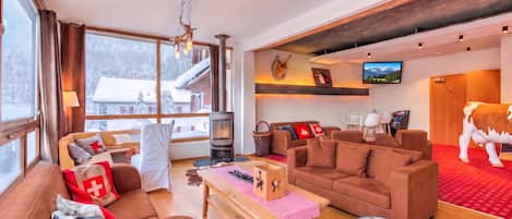 A spacious living room with a fireplace, comfortable seating, a television, and a large window with a view of a snowy landscape.