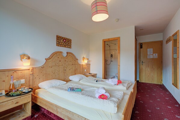 Une chambre d’hôtel avec deux lits, des têtes de lit en bois, une table de chevet en bois avec une lampe, une petite table avec une bouteille et des verres, et une moquette rouge.