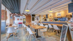 Een modern restaurantinterieur met lange gedeelde tafels en witte stoelen, een houten toonbank en een menukaart op een schoolbord.