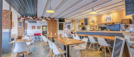 A modern restaurant interior with long communal tables and white chairs, a wooden counter, and a chalkboard menu.
