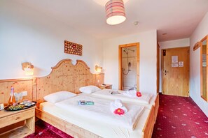 Une chambre d’hôtel avec une tête de lit en bois, un lit recouvert de draps blancs, une table de chevet avec une bouteille et des verres, un espace douche et un sol recouvert d’une moquette rouge.