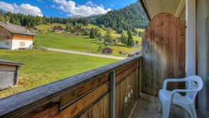 Ein Balkon mit Holzwertkasten, einem weißen Plastikstuhl und Blick auf ein grünes Tal mit Häusern und Bäumen unter blauem Himmel.