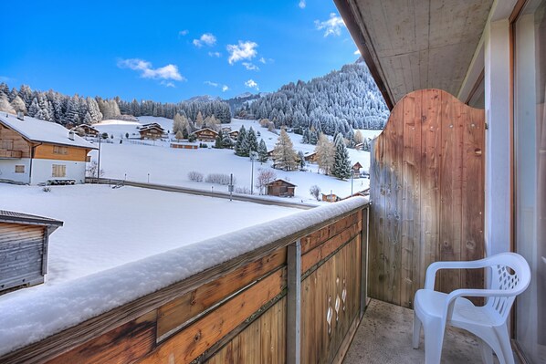 A balcony with a wooden railing, a white plastic chair, and a snowy landscape with houses in the background. - Hotel Relais Alpin Twin Room & Balcony 3 136 summer mode, Les Mosses, Switzerland (Les Mosses)