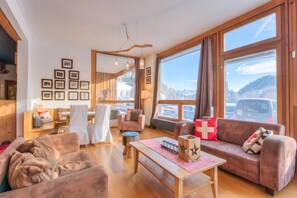 A cozy living room with a sofa, chairs, a coffee table, and a fireplace. Large windows offer a view of the outdoors. - Hotel Relais Alpin Twin Room & Balcony 3 136 summer mode, Les Mosses, Switzerland (Les Mosses)