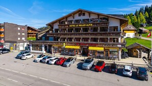 Un hotel con balcón, restaurante y aparcamiento con varios coches.