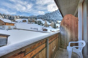 Un balcón con barandilla de madera, una silla de plástico blanca y un paisaje nevado con casas al fondo.