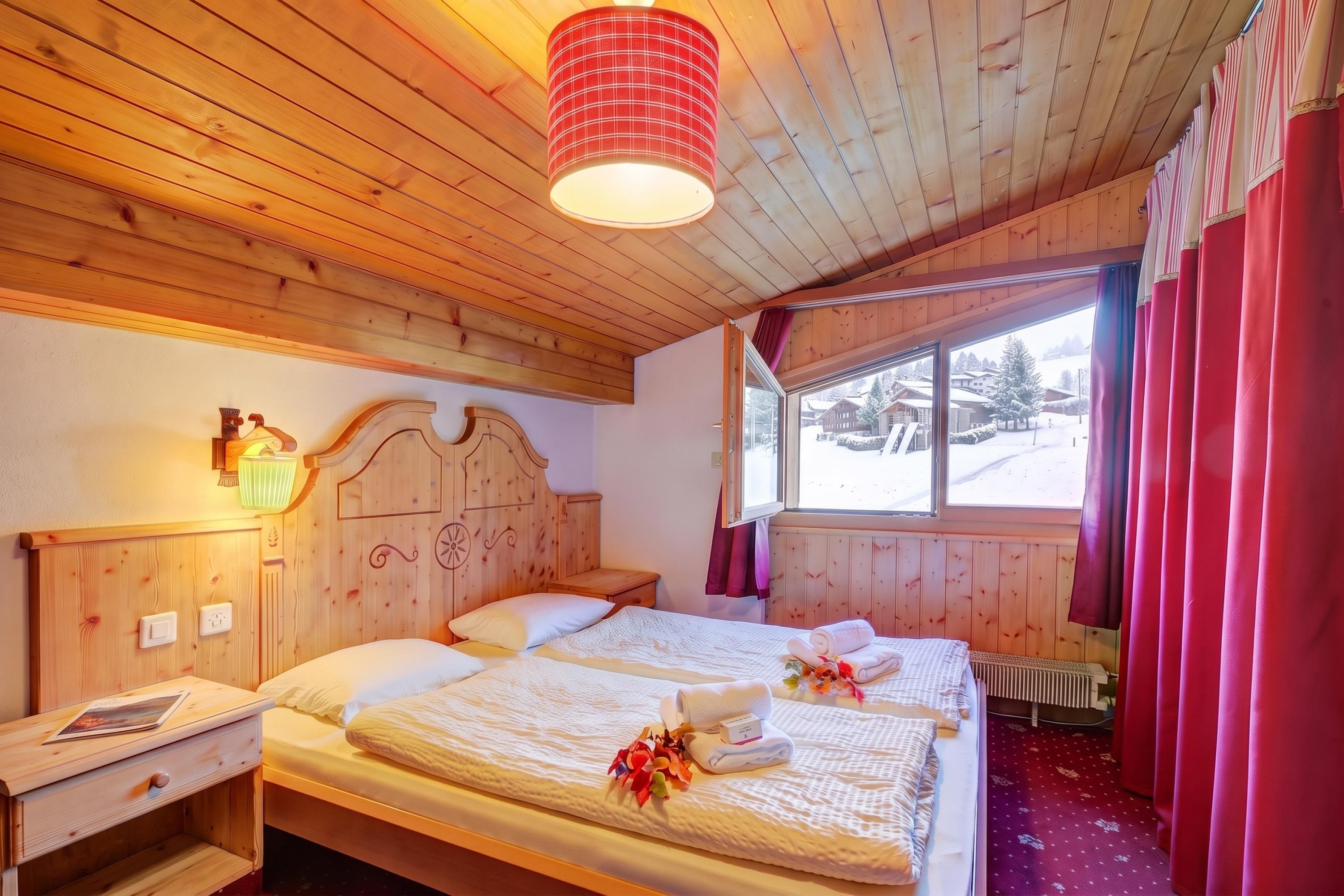 Habitación de cabaña de madera con dos camas, una ventana con vistas a un paisaje nevado y una lámpara roja.