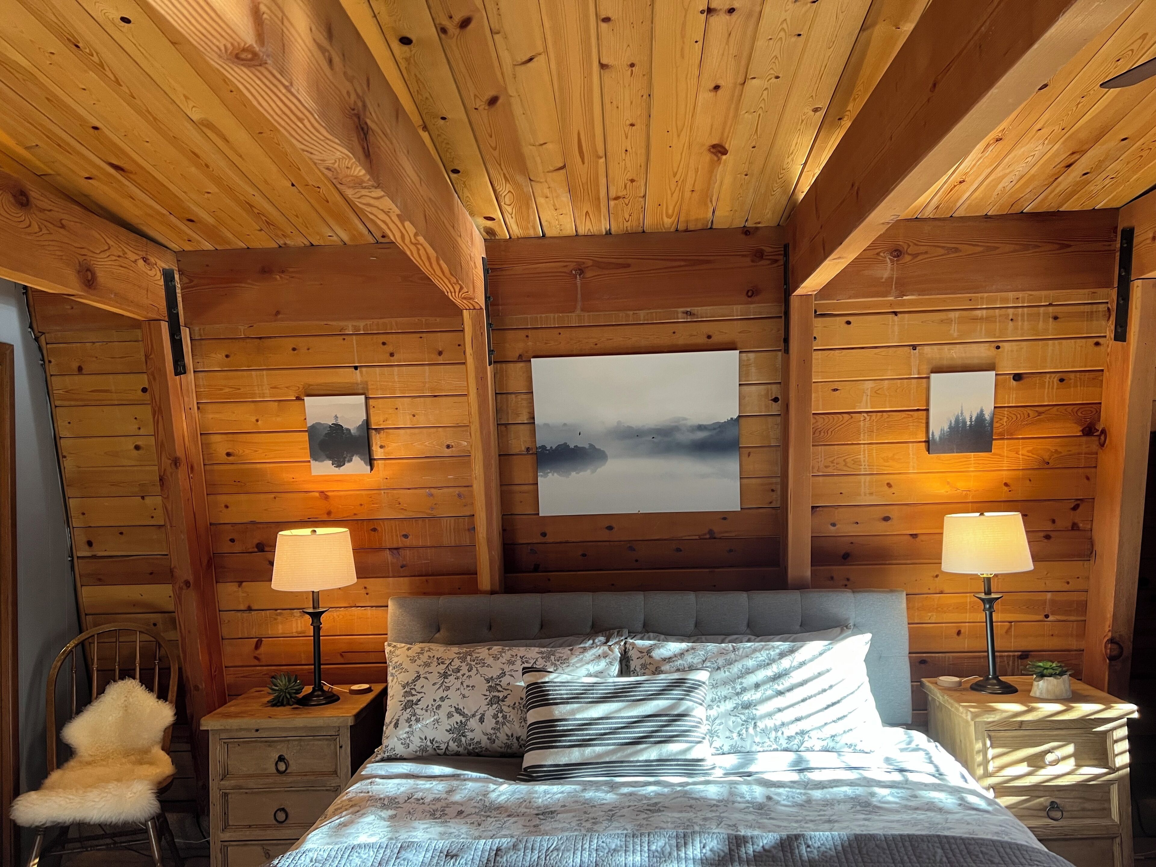 Ein Schlafzimmer einer Holzhütte mit einem Bett, zwei Nachttischen, einem Sessel und Kunstwerken an den Wänden.
