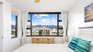 A bedroom with a large window, a bed, a wooden dresser, and a view of a cityscape. - Waikiki Banyan condo w/ pool & beach access (Honolulu)